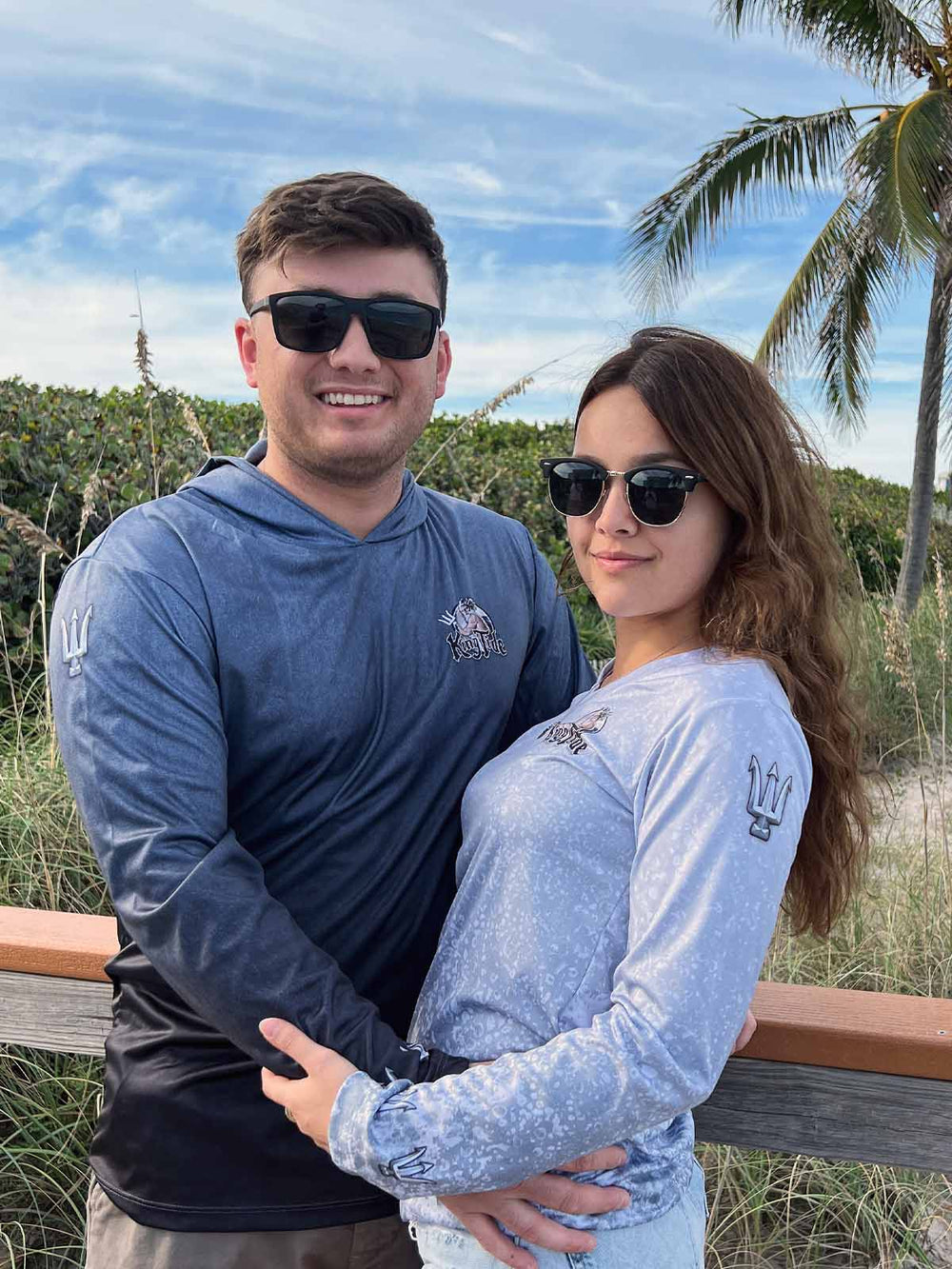 Couple standing on a beach. He is wearing King Tide serene black hoodie. She is wearing a slate fossil print long sleeve performance shirt.