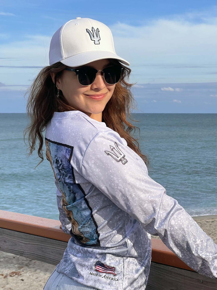 Long Sleeve, V neck performance shirt with slate fossil allover print. Right side with trident on sleeve at shoulder. American flag 'Made in America' on bottom / back. Model is also wearing a King Tide white hat with titan chrome 3D trident logo.